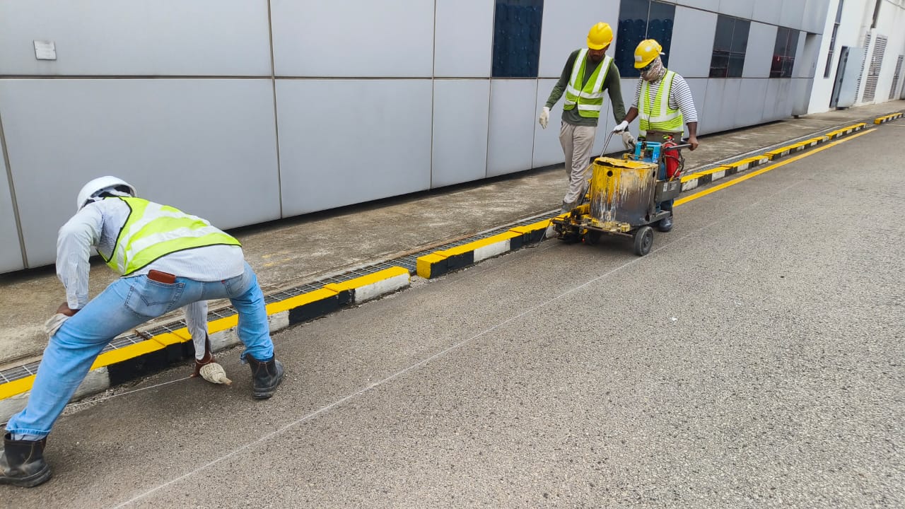 ASU crew operating thermoplastic line-marking machine along yellow safety curb on industrial driveway